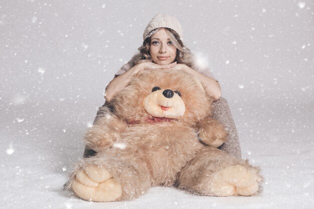 Casual smiling young woman in knitted clothes holding big soft teddy bear on snowy background