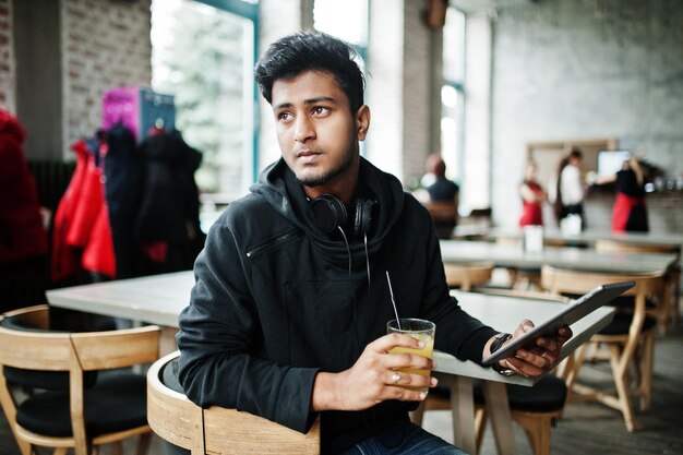 Вскользь и стильный молодой азиатский человек с наушниками на кафе выпивая сок от стекла и держа таблетку