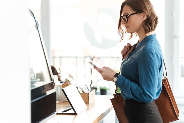 casual woman in eyeglasses standing with mobile phone indoors