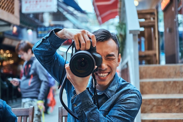 Casual young asian men is making a photo with photo camera outside in public place. He is smiling. Man is wearing denim jacket.