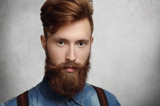 casual young bearded man with stylish haircut and freckles posing against blank concrete wall with copy space for text or advertising content, looking serious.