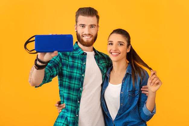 Casual young couple holding wireless speaker happy listening to music dancing colorful style happy mood isolated on yellow wall