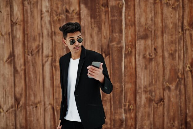 Casual young indian man in black blazer and sunglasses posed against wooden background looking at mobile phone