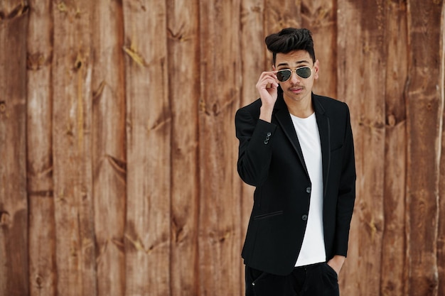 Casual young indian man in black blazer and sunglasses posed against wooden background