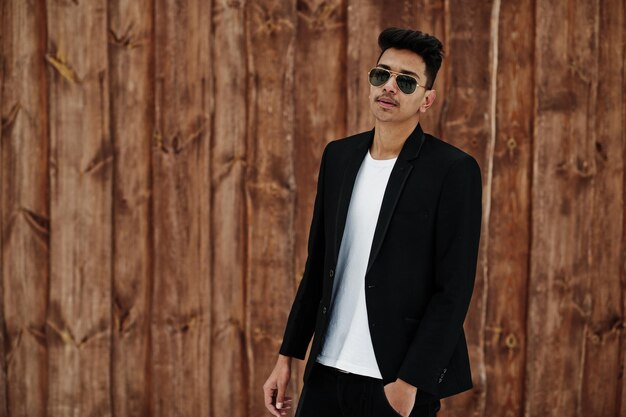 Casual young indian man in black blazer and sunglasses posed against wooden background