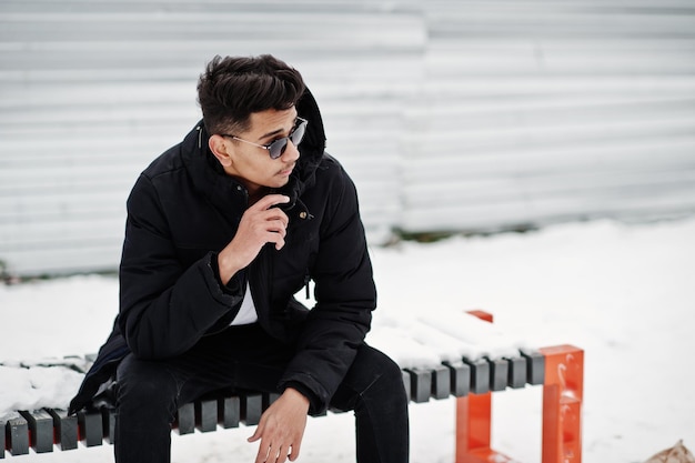 Casual young indian man in black jacket and sunglasses posed at winter day and sitting on bench