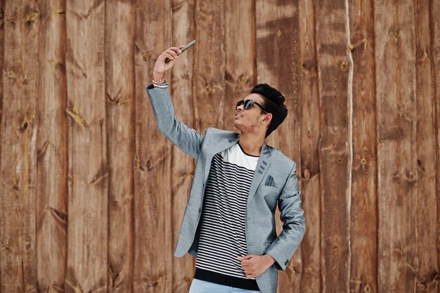 Casual young indian man in silver blazer and sunglasses posed against wooden background making selfie on phone