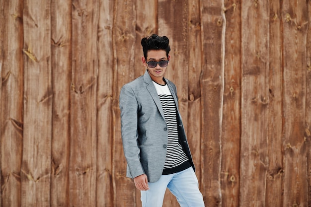 Casual young indian man in silver blazer and sunglasses posed against wooden background