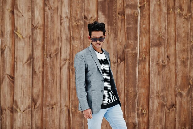 Casual young indian man in silver blazer and sunglasses posed against wooden background