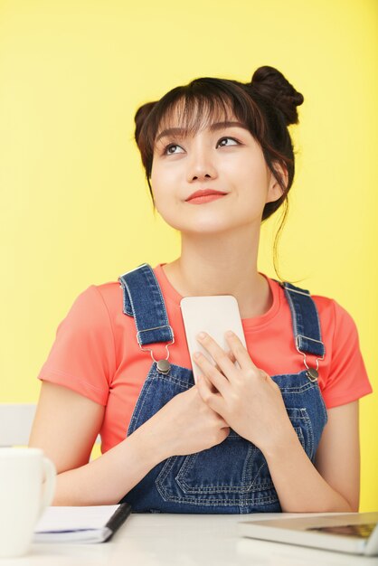 Casually dressed Asian woman sitting at desk, looking up and clasping smartphone to chest 