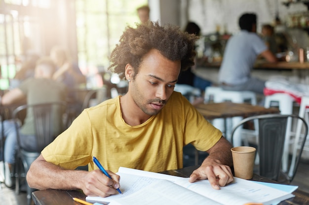 Casually dressed young black male student with beard and curly hair having focused concentrated look as he reads information in textbook and making notes in copybook, preparing for lesson at college