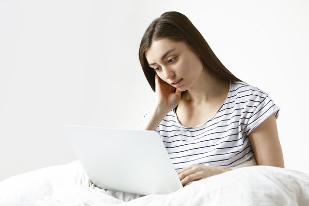 Небрежно одетая молодая европейская женщина-фрилансер использует обычный ноутбук для удаленной работы, сидит на белой кровати и смотрит на экран