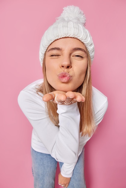 Catch my kiss Romantic woman keeps lips rounded sends air kiss keeps palm forward camera near folded lips stands coquettish wears winnter hat and white turtleneck isolated over pink background