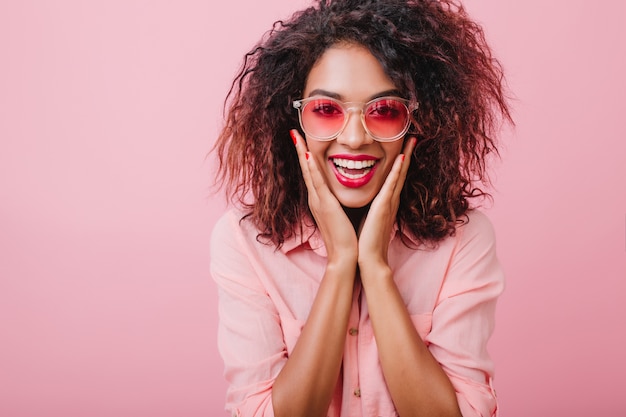 Cattura ragazza riccia in occhiali da sole alla moda agghiacciante e ridendo. adorabile donna dai capelli scuri in abiti di cotone rosa in posa con la faccia sorpresa.