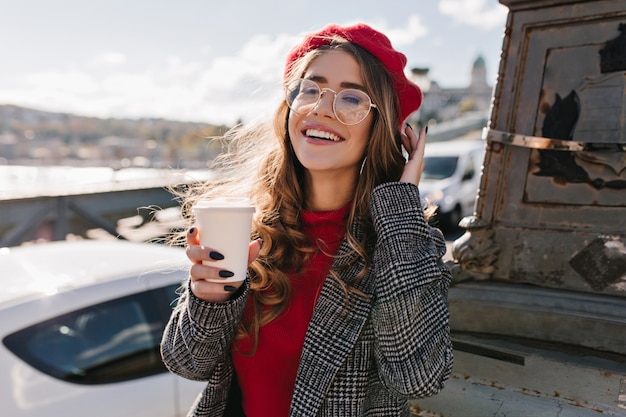 Foto gratuita ragazza di cattura con l'espressione del viso eccitato che beve caffè per strada in una giornata ventosa e fredda