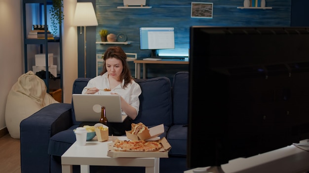 Caucasian adult eating chinese food while using laptop at home in living room. Woman holding asian meal with chopsticks in hand watching television and working on modern device on couch