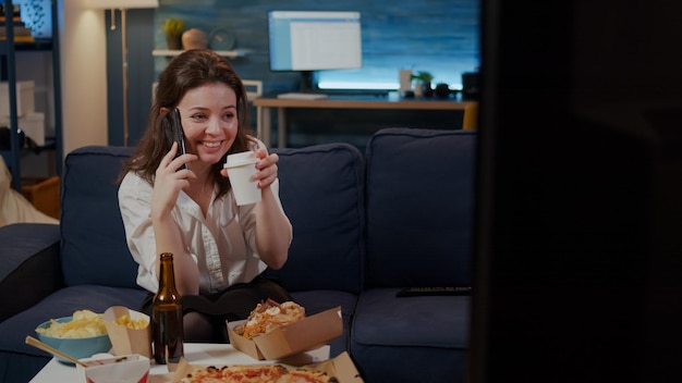 Caucasian adult enjoying phone call with friend in living room while drinking coffee. Woman talking on smartphone, preparing to eat fast food delivery meal at home after work to relax