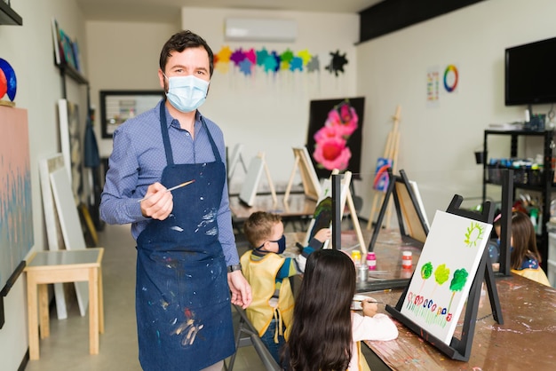 Caucasian adult man with a face mask and an apron during an art class for kids. Male teacher instructing two kids how to do a painting