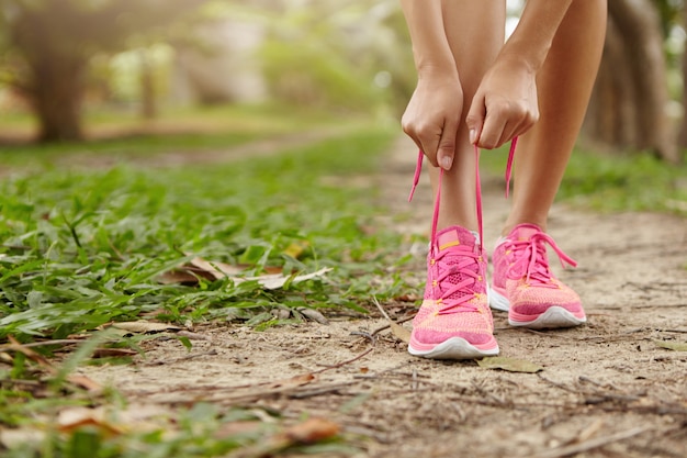 Donna atletica caucasica che lega i lacci sulle sue scarpe da corsa rosa prima di fare jogging in piedi sul sentiero nella foresta. corridore femminile che allaccia le sue scarpe da ginnastica mentre fa l'allenamento nella zona rurale.