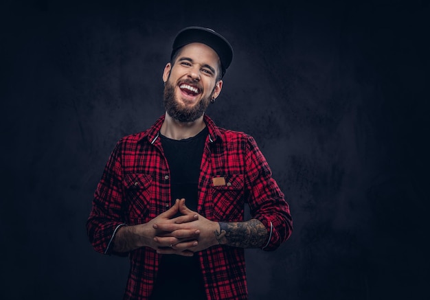 A caucasian bearded hipster male laughing dressed in a flannel shirt and a cap, posing in studio, over dark background.