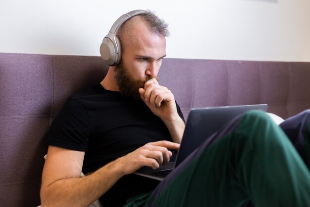 Caucasian bearded man in headphones in bedroom on bed working on laptop from home, typing, thinking.