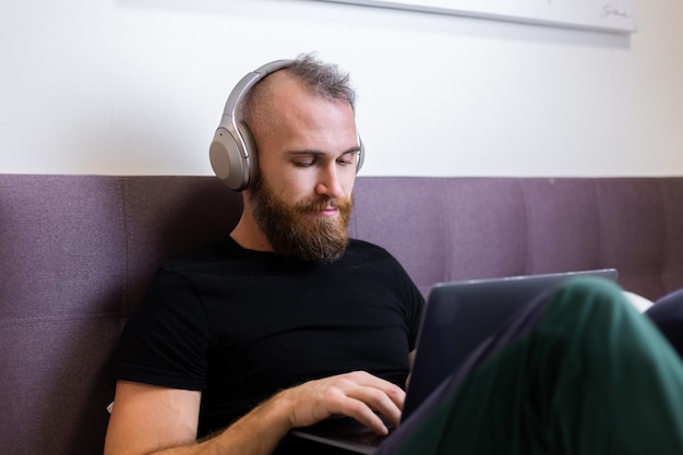 Caucasian bearded man in headphones in bedroom on bed working on laptop from home, typing, thinking.