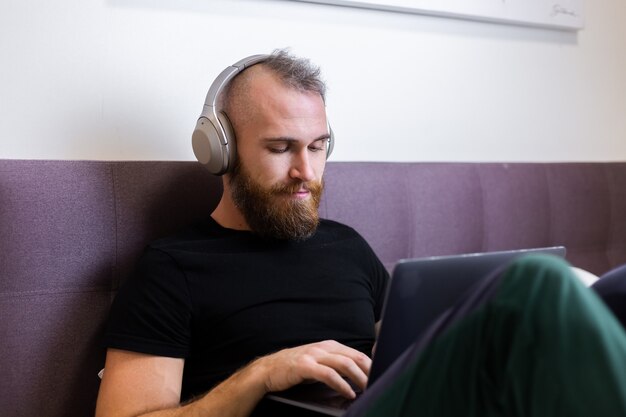 Caucasian bearded man in headphones in bedroom on bed working on laptop from home, typing, thinking.