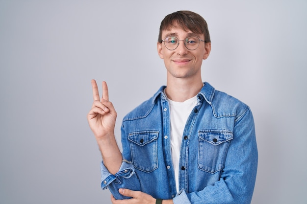 Foto gratuita uomo biondo caucasico in piedi con gli occhiali sorridente con faccia felice strizzando l'occhio alla telecamera facendo segno di vittoria con le dita. numero due.