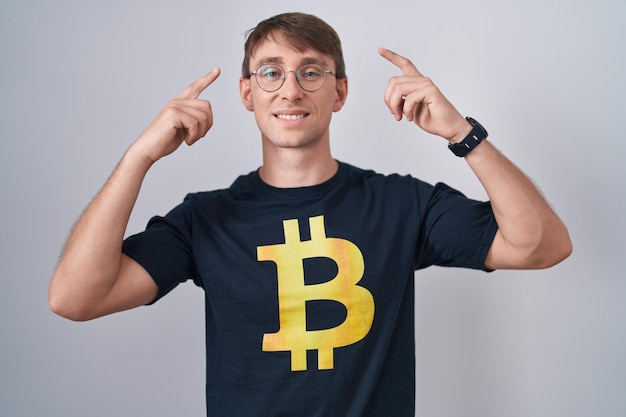 Free photo caucasian blond man wearing bitcoin t shirt smiling pointing to head with both hands finger, great idea or thought, good memory