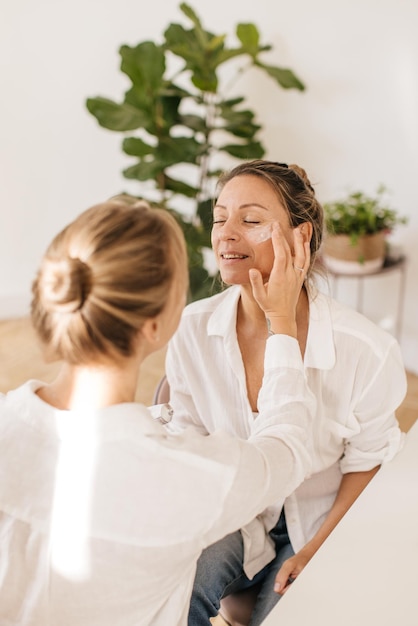 Caucasian blonde girl applies anti-aging cream for skin aging on her mother's face indoors. Concept of natural cosmetics, wrinkle smoothing