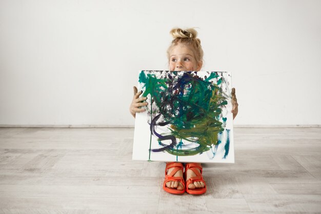 Caucasian blonde preschool girl showing picture that she painted. Adorable child holding canvas. Happy childhood concept.