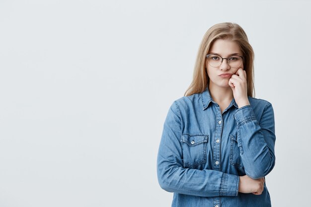 Caucasian blonde pretty female in denim shirt wearing glasses pouting her lips in dissatisfaction while doubting. Thoughtful girl crossing arms looks with disappointment and offense 