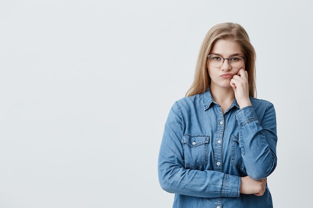 Caucasian blonde pretty female in denim shirt wearing glasses pouting her lips in dissatisfaction while doubting. Thoughtful girl crossing arms looks with disappointment and offense 