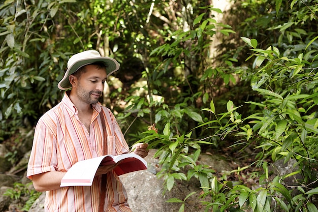 Caucasian botanist or biologist with stuble wearing striped shirt and hat holding notebook in one hand and green leaf of exotic plant in another having joyful expression on his face, enjoying his work