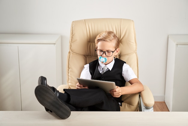 Caucasian boy sitting in executive chair in office with dummy in mouth and using tablet