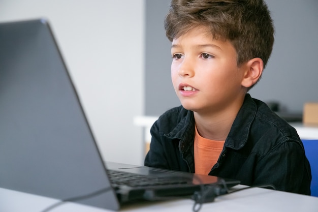 Caucasian boy sitting at table in classroom, reading text on screen or watching video presentation