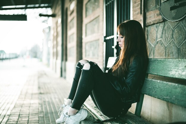 Caucasian brunette female sitting on the bench behind a building and waiting for the train