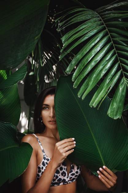 Caucasian brunette woman with golden tan hiding behind tropical leaves and palm trees standing in summer bikini and looking sensual Concept of tourism and tropic holidays