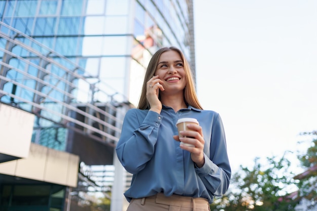 行くコーヒーを保持している電話で話す白人のビジネス女性。電話で話して、近代的なオフィスビルの上に立って成功したヨーロッパの女性