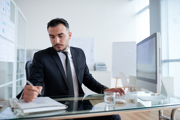 Free photo caucasian businessman sitting at desk in front of computer and writing on document folder