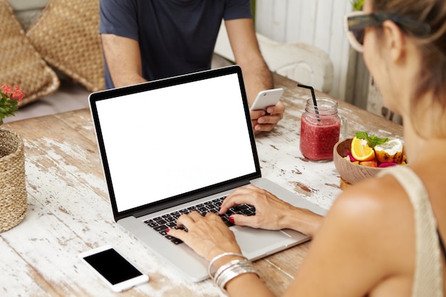 カフェで無料のwi-fiを使用し、電子機器でオンライン通信を楽しんでいる白人のカップル：夫が携帯電話でSMSをテキストメッセージしている間にラップトップコンピューターで電子メールをチェックしている女性