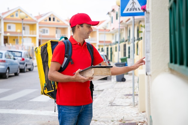 Caucasian courier carrying package and ringing on doorbell while making home delivery. Middle-aged deliveryman with yellow thermo bag standing in front of house. Delivery service and post concept