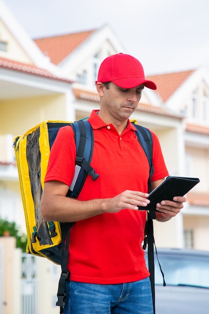 Free photo caucasian courier standing and reading address in tablet. focused deliveryman delivering express order in yellow thermal backpack.