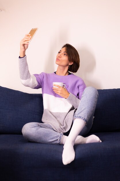 Кавказская милая женщина-блогер дома в теплом фиолетовом свитере делает селфи в зеркале на мобильном телефоне