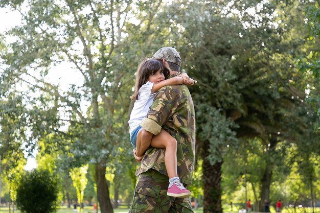 Caucasian dad in army uniform embracing daughter. Middle-aged father standing in city park. Cute girl sitting on his hands and hugging daddy on neck. Childhood, weekend and military parent concept
