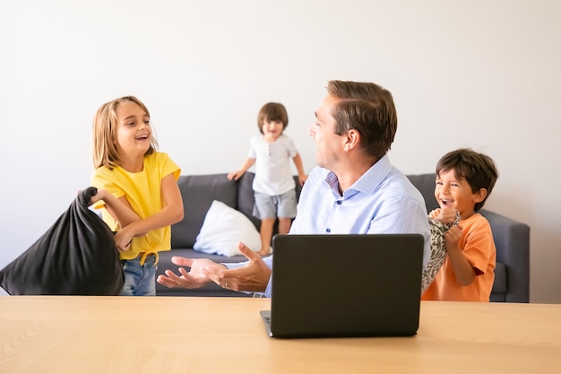 Caucasian dad talking with playful kids and sitting at table. Happy middle-aged father using laptop computer when children playing with pillow at home. Childhood and digital technology concept