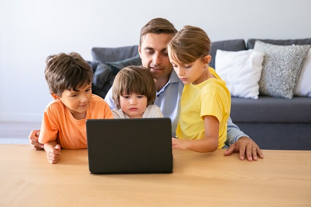 Caucasian dad watching movie via laptop with kids. Happy father sitting at table with lovely children. Cute thoughtful boys and blonde girl looking at screen. Childhood and digital technology concept