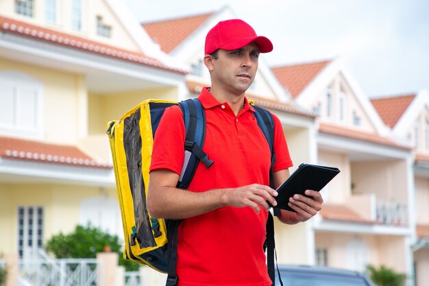 Caucasian deliveryman looking for address and holding tablet. Thoughtful courier delivering order in yellow thermo backpack.