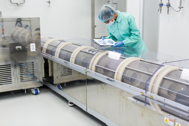 Caucasian factory worker in blue lab suit with tablet in hands bend on manufacture machine check readings