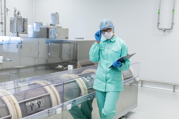 Кавказский фабричный рабочий в синем лабораторном костюме с планшетом в руках стоит возле производственной машины, рука на очках